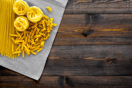 Assortment of raw pasta. Spaghetti, fusilli, penne, fettuccine on blue tablecloth on dark wooden background top view.の写真素材