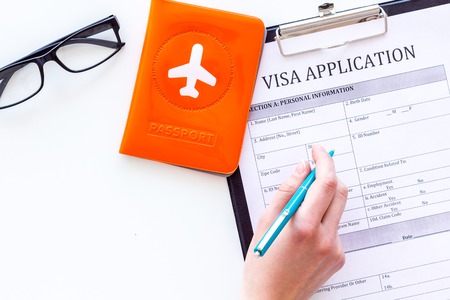 Hand fills visa application form. Form near glasses, pen, passport cover with airplane sign on white background top view.の写真素材