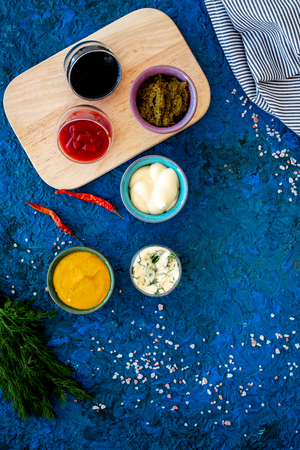Spicy sauses. Ketchup, mayonnaise, mustard, soy sauce, barbecue sauce, pesto, mustard, sour near chili pepper and greenery on blue background top view copy spaceの写真素材