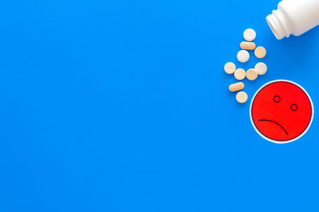 Illness. Drug intake. Pills falling out of jar near sad face emoji on blue background top view copy spaceの写真素材