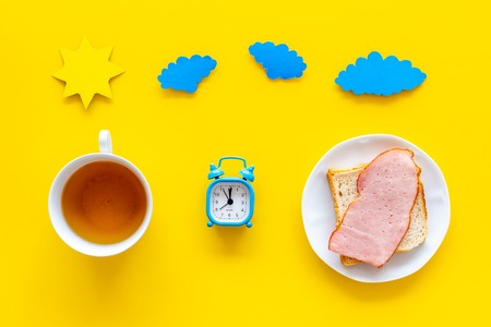 Parts of day. Morning. Time for breakfast. Tea, sandwich near alarm clock, sun and clouds cutout on yellow background top view.の写真素材