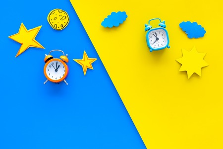 Sleep time, clock on the bed and time to awake concept. Alarm clock near sun, moon, stars cutout on blue and yellow background top view.の写真素材