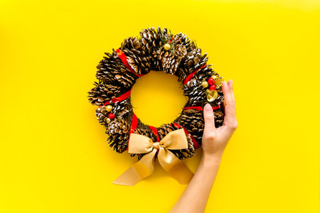 Hand hang a christmas wreath made of pine cones on yellow background top view.の写真素材