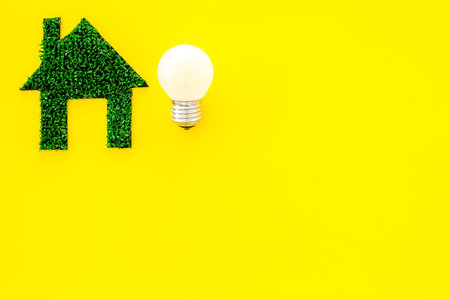 Enegry saving technology concept. House cutout made of green grass near light bulb on yellow...