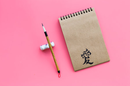 Chinese traditional calligraphy concept. Asian hieroglyph love in craft paper notebook near special writing pen on pink background top view copy spaceの写真素材