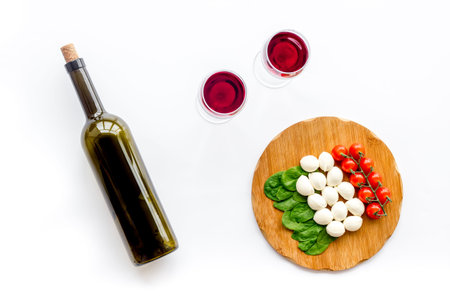 Italian cuisine, food concept. Italian flag made of mozzarella, tomatoes, basil on wooden cutting board near glass of red wine and wine bottle on white background top viewの写真素材