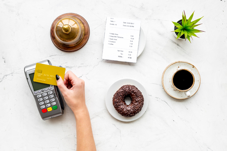 Pay the bill by payment terminal. Womans hand insert bank card in payment terminal near bill, service bell, coffee on white stone background top viewの写真素材