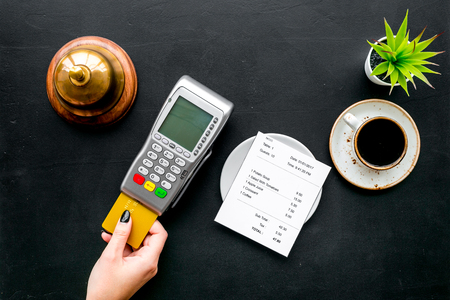 Pay the bill by payment terminal. Woman's hand insert bank card in payment terminal near bill, service bell, coffee on black background top view.の写真素材