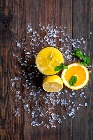 Juice in glass near cut orange and lemon, crushed ice, green mint on dark wooden background top view.の写真素材