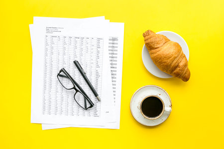 Breakfast in office with croissant and black coffee on the work desk with documents and glasses on yellow background top viewの写真素材