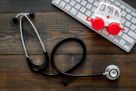 Call doctor online concept. Ambulance vehicle toy near computer keyboard and stethoscope on dark wooden background top viewの写真素材