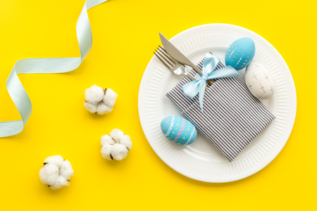 Easter table decoration. Plate, cutlery, painted eggs and dry white flowers, tablecloth on yellow background top view.の写真素材