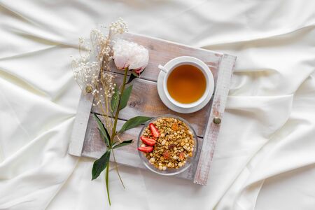 Granola with fruit, cup of tea and flower on tray for breakfast in bed on white sheets background top viewの写真素材