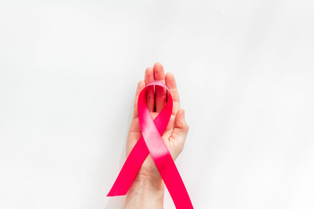 Pink ribbon in hands as symbol of breast cancer awareness on white background top view space for...