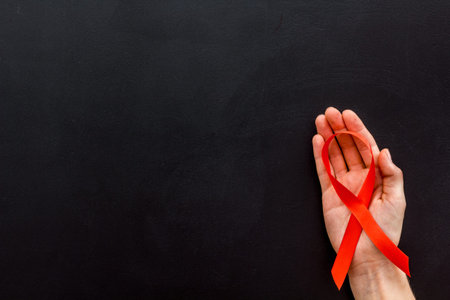 AIDS disease symbol. Red ribbon in hands on black background top view.