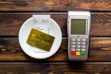Electronic payments in restaurant. Bank card near aquiring terminal and bill on dark wooden...