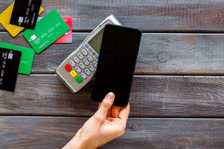 Contactless payment, NFC technology. Hand hold phone near terminal on dark wooden background...