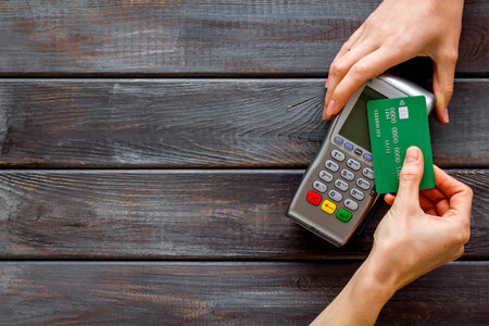 Contactless payment, paypass. Hand hold bank card near terminal on dark wooden background top-down...