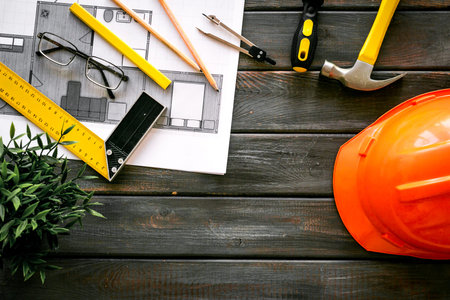 Builder work desk with hard hat, instruments and blueprints on dark wooden background top-down.