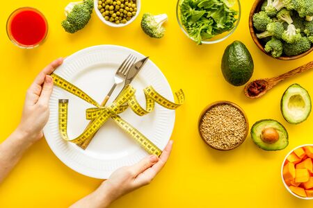 Diet concept. Fork and knife tied with measure tape on plate in hands on yellow background with vegetables top view.の写真素材