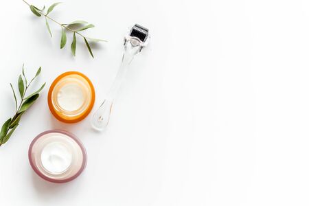 Cosmetologist tools. Dermaroller, creams near leaves and flowers on white background top-down frame copy spaceの写真素材