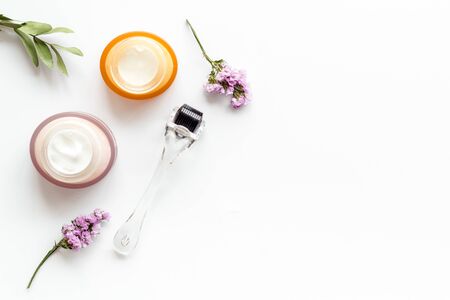 Cosmetologist tools. Dermaroller, creams near leaves and flowers on white background top-down frame copy spaceの写真素材