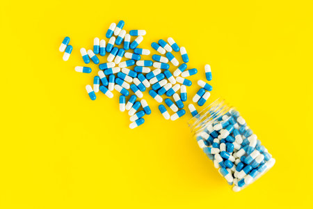 Blue and white capsules poured from a bottle - sports supplement creatine - on yellow background top...