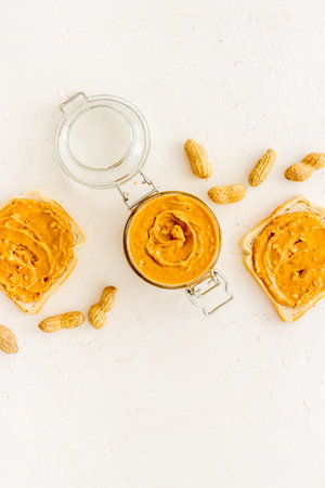 Glass jar of peanut butter with sandwiches and nuts on kitchen table