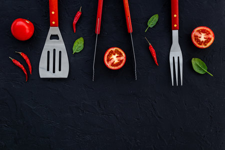 Picnic flatlay. Steel barbecue utensils with tomatoes and greens, top view.の写真素材