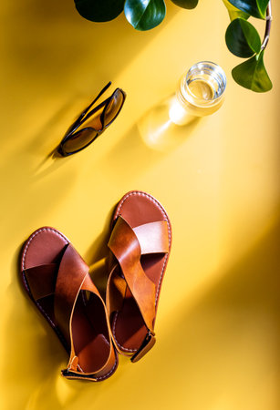 Summer sunlight shadows on sandals, sunglasses and a glass of water on yellow background, top view. Beach concept.の写真素材
