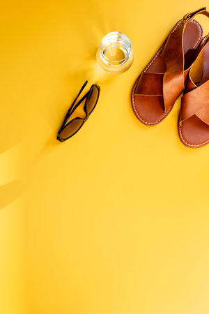Summer sunlight shadows on sandals, sunglasses and a glass of water on yellow background, top view. Beach conceptの写真素材