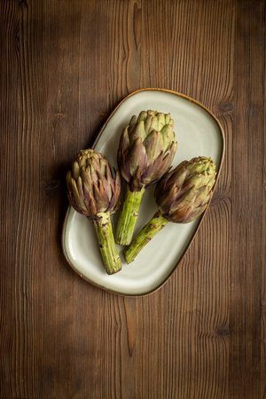 Three artichokes flowers - purple edible buds on a plate top view. Vegetarian food ready to cookの写真素材