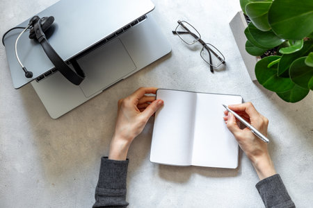 Hands writing in notebook near headset and laptop, top view. Customer support equipment at call center. Online conference. Online education concept.の写真素材