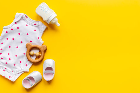 Baby bodysuits and eco friendly accessories - booties, wooden rattle toy and baby bottle with milk on yellow background, top view.の写真素材