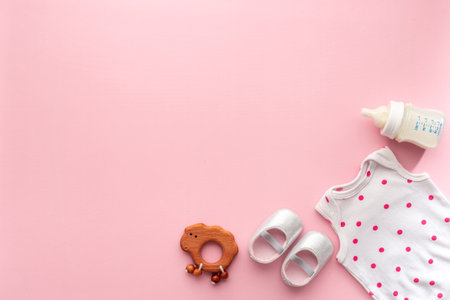 Flat lay of baby bodysuits and eco friendly accessories - booties, wooden rattle toy and baby bottle with milk on pink background, top view.の写真素材