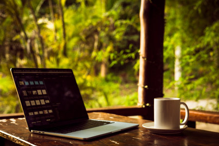 Laptop and cup on wooden table of terrace with tropical forest view at sunset, remote work digital nomad.の写真素材