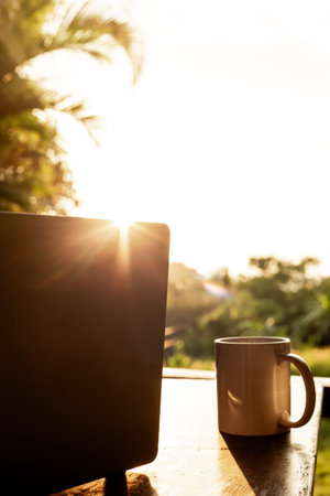 Laptop and cup on wooden table with forest view at sunset, remote freelance work. Sunshine and backlight.の写真素材