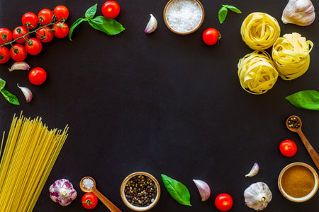 Frame of ingredients for cooking Italian pasta - uncooked spaghetti with tomatoes and basil leaves - on a black background, top view.の写真素材