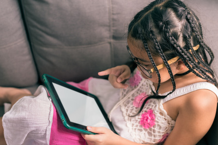 Cute little girl,wearing glasses,dreadlocks hair style ,playing with computer at home laying on sofaの写真素材