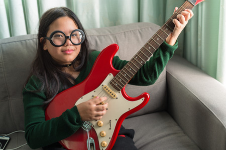 Happy smiling girl ,wearing glasses,learning to play the Electric guitar at home laying on sofaの写真素材