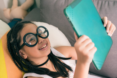 Cute little girl,wearing glasses,dreadlocks hair style ,playing with computer at home laying on sofaの写真素材