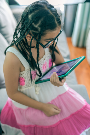 Cute little girl,wearing glasses,dreadlocks hair style ,playing with computer at home laying on sofaの写真素材