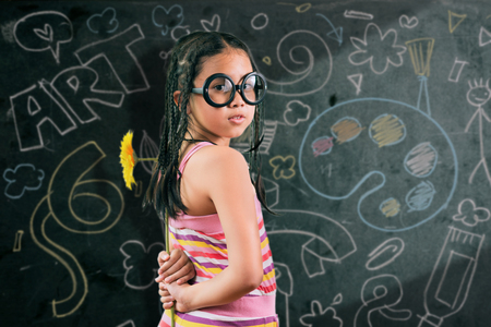 Portrait of cute girl in eyeglasses looking at camera by the blackboard with chalk drawings of Artの写真素材
