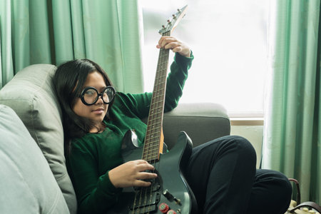 Happy smiling girl ,wearing glasses,learning to play the Bass guitar at home laying on sofaの写真素材