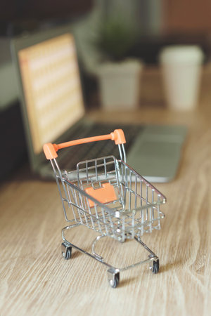 Online shopping concept, Shopping cart with blurry laptop on the desk