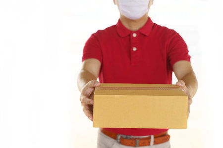 delivery man with face mask in red cloth holding a box package. Isolated on white studio Backgroundの写真素材