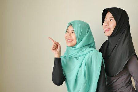 happy muslim woman, smiling islamic women pointing up; muslim girls point up; beautiful asian muslim women with hijab or head scarf pointing finger or pointing hand to blank spaceの写真素材