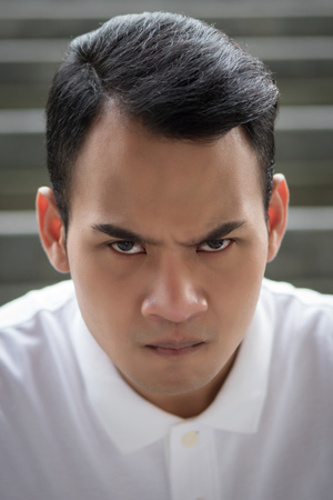 unhappy man portrait, angry man face, hostile handsome man sitting looking at you with bad angry mood emotion; young adult south east asian man model with tan skin and short hairの写真素材