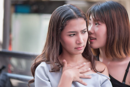 unhappy woman gossip with bad news; serious, frustrated, unhappy women whispering, spreading gossip or bad news to her friend, concept of mouth information or news transmission; asian 20s woman modelの写真素材