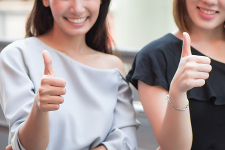 successful girl pointing thumb up for success; two women giving thumb up to you; portrait of cheerful smiling women pointing up approving, yes, ok, good, thumb up gesture; asian woman 20s adult modelの写真素材